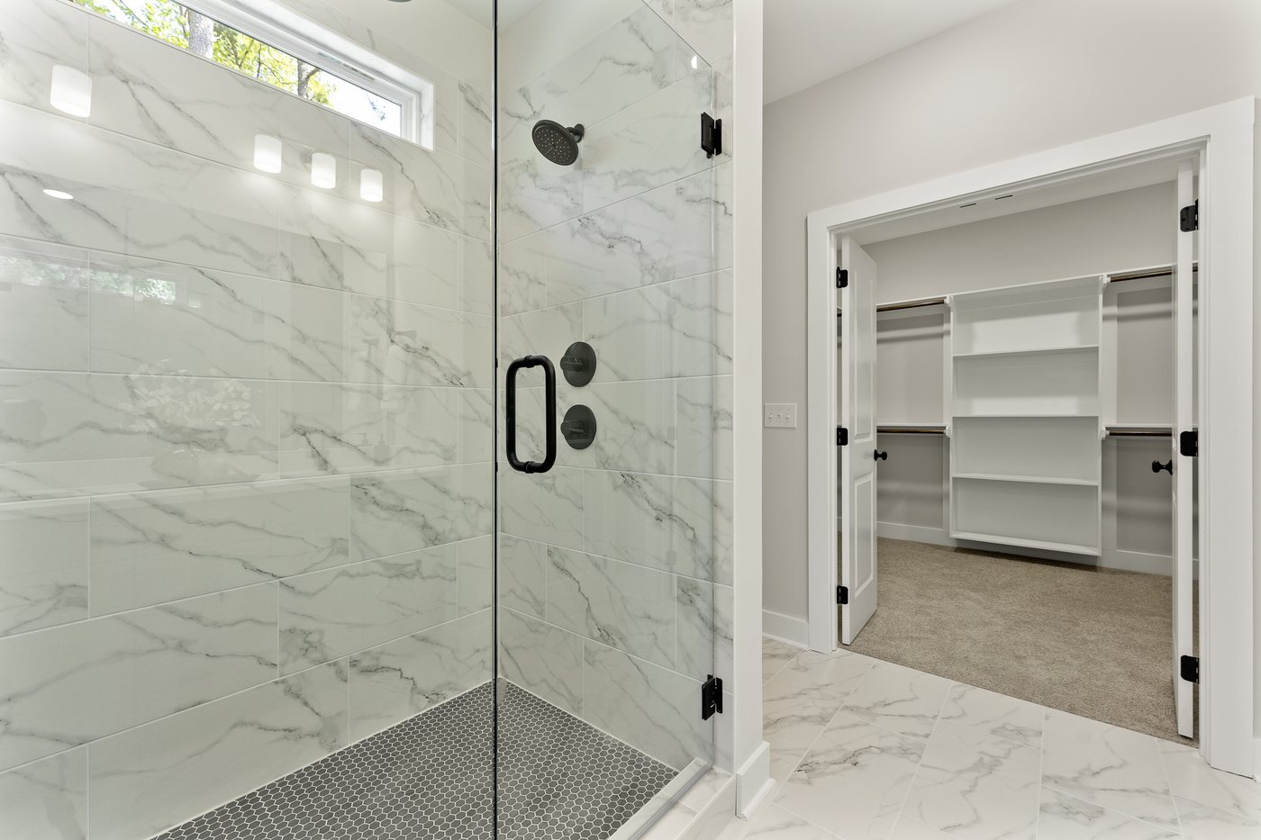 A finished Hornet primary bathroom with a marble-look tile walk-in shower, black fixtures, and a built-in walk-in closet beyond.